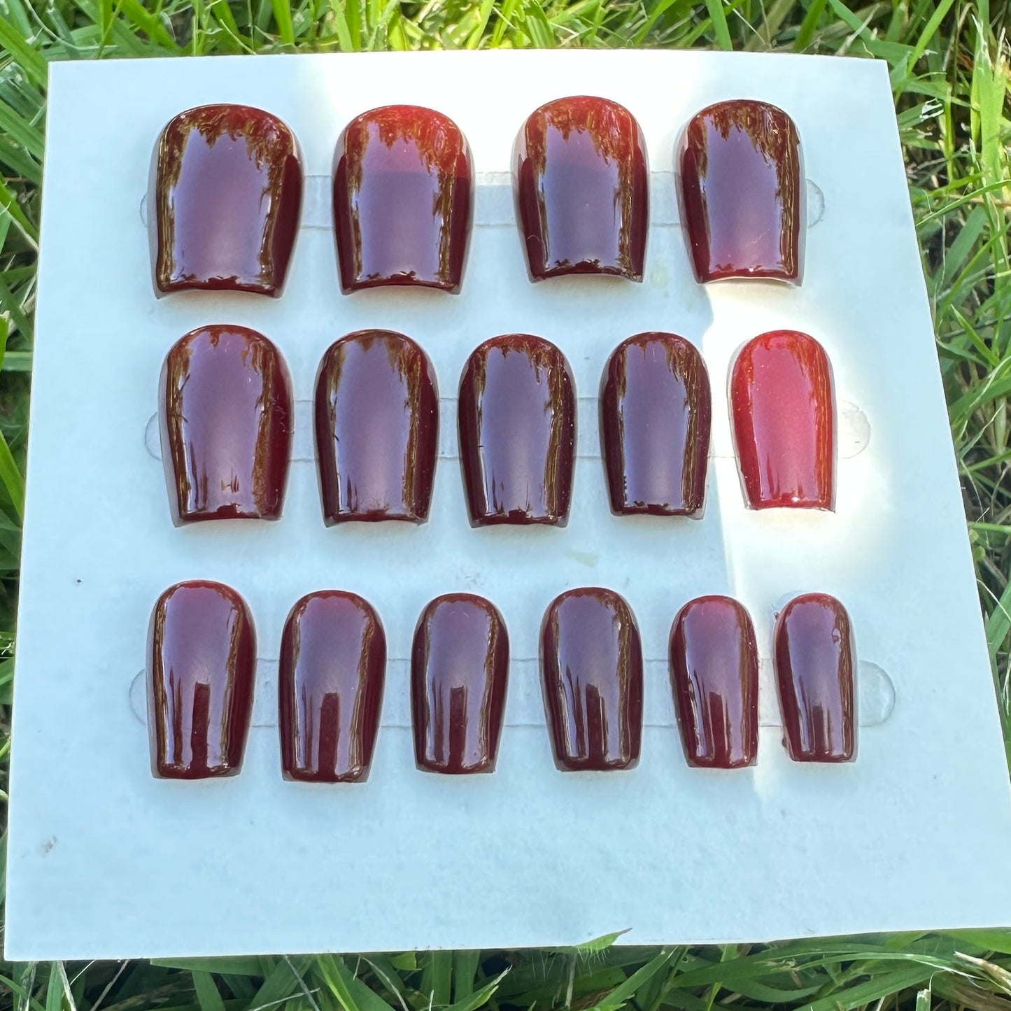 Set of red nail polish samples on a white card with grass in the background. Red nail theory inspired nails on hand model