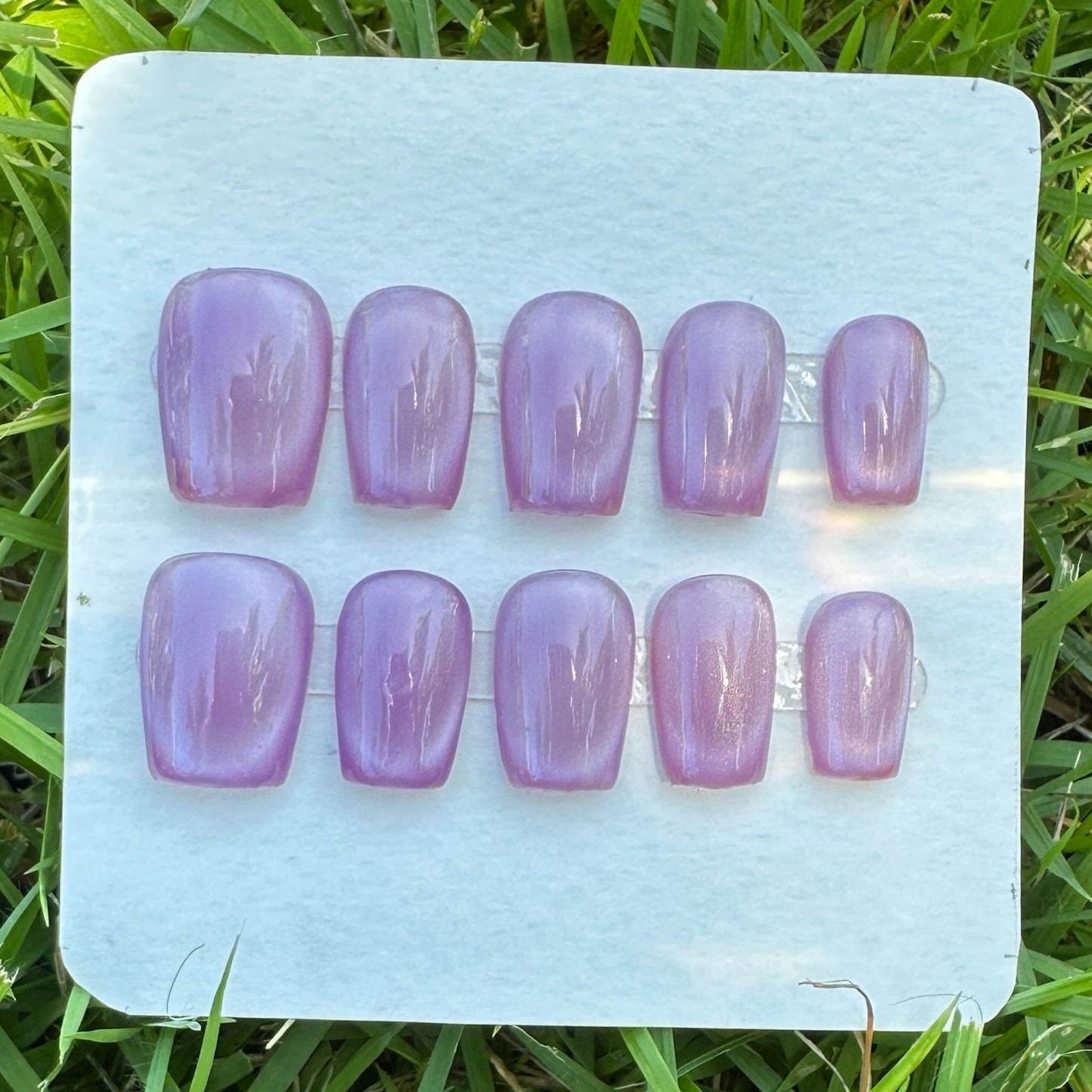 Set of purple nail extensions on a clear plastic tray placed on grass. purple full moon cat eyes. 