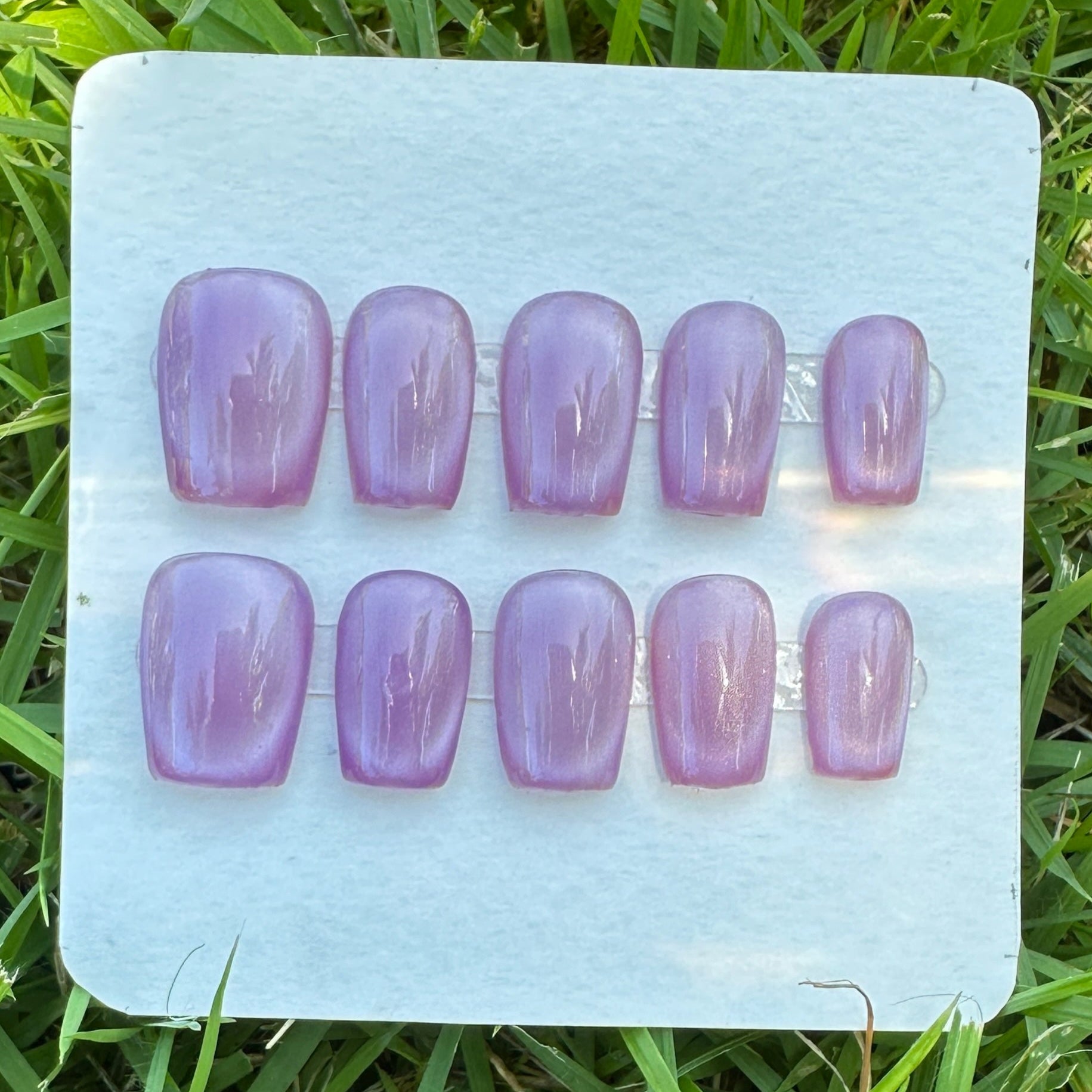 Set of purple nail extensions on a clear plastic tray placed on grass. purple full moon cat eyes. 